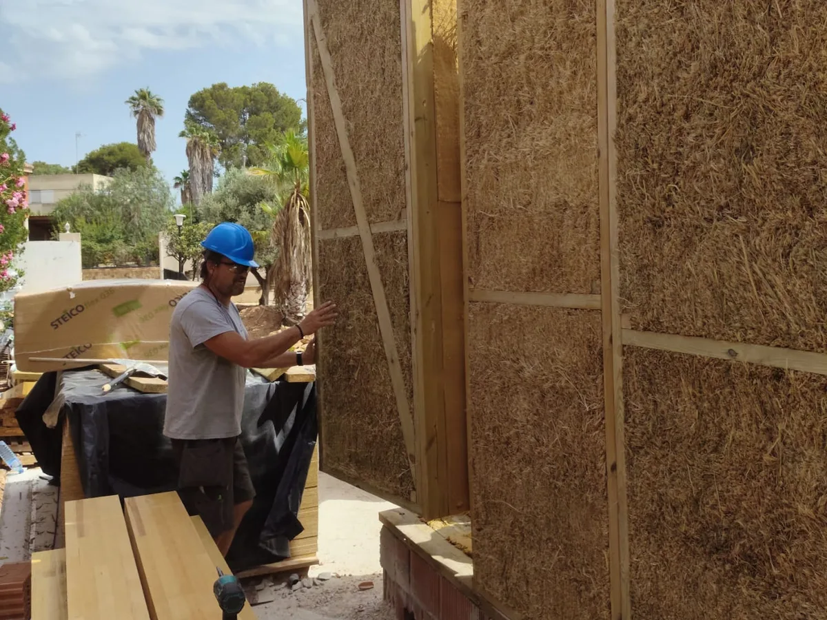 Operario guiando un panel de madera relleno de paja mientras la grúa lo coloca sobre la base de ladrillo